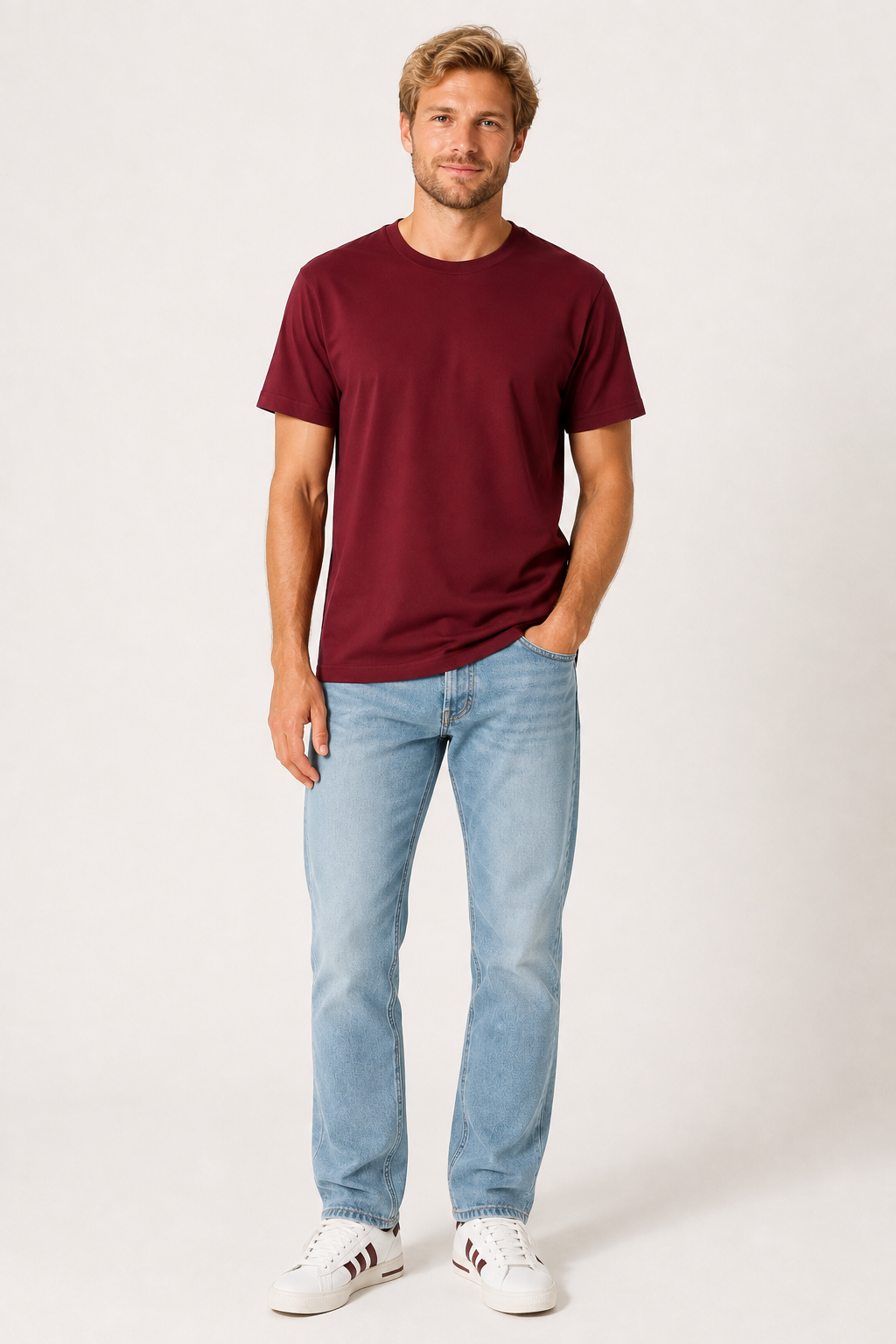 A man modeling a short-sleeve crewneck t-shirt in burgundy, paired with light blue jeans and white sneakers, standing against a plain off-white studio background.