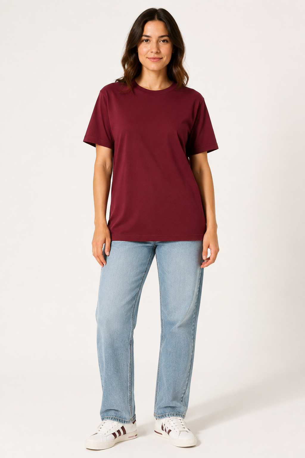 A woman modeling a short-sleeve crewneck t-shirt in burgundy, paired with light-wash blue jeans and white sneakers, standing against a plain light gray studio background.